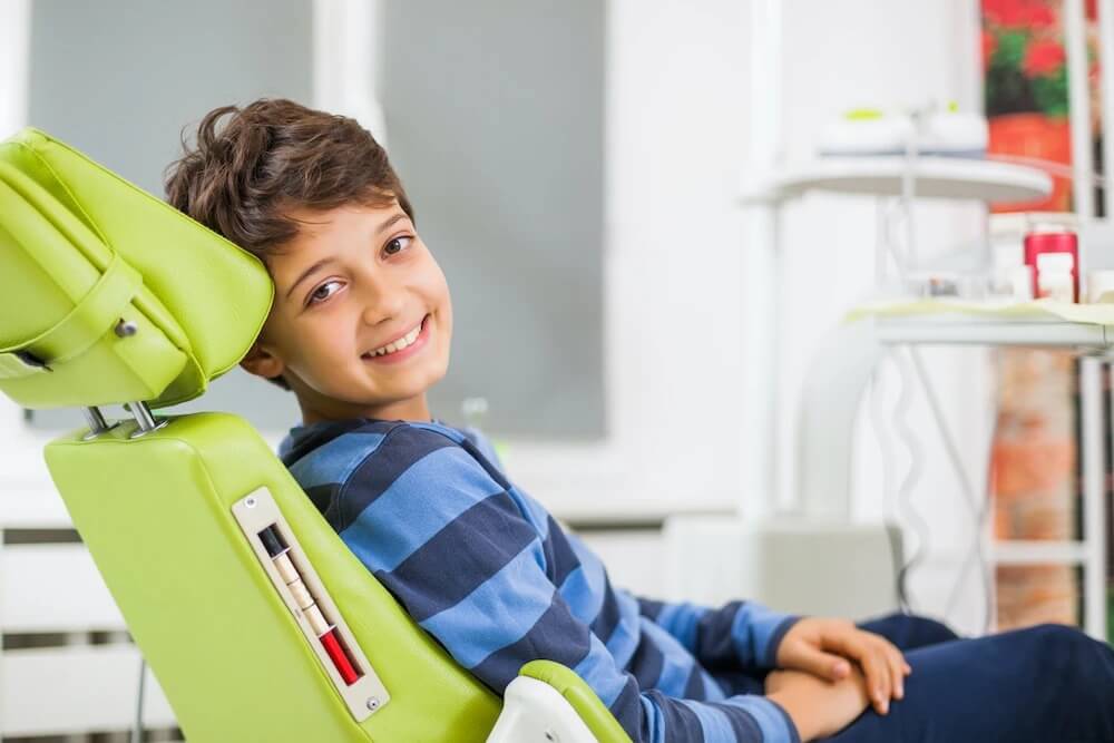 Brunette boy in a striped shirt smiles while sitting in a green dental chair at Safari Children's Dentistry & Braces in San Antonio, TX