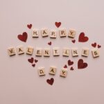 Scrabble letters spell out Happy Valentine's Day surrounded by red sticker hearts on a pink background